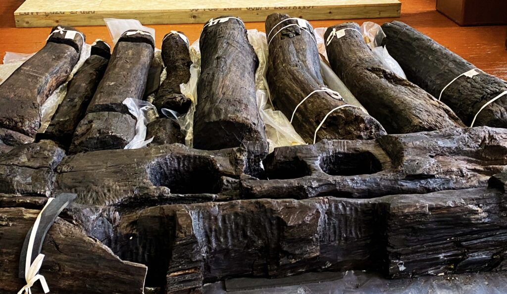 Heavy dark timbers laid in rows, some with large notches and mortices, tool marks showing they have been deliberately shaped. 